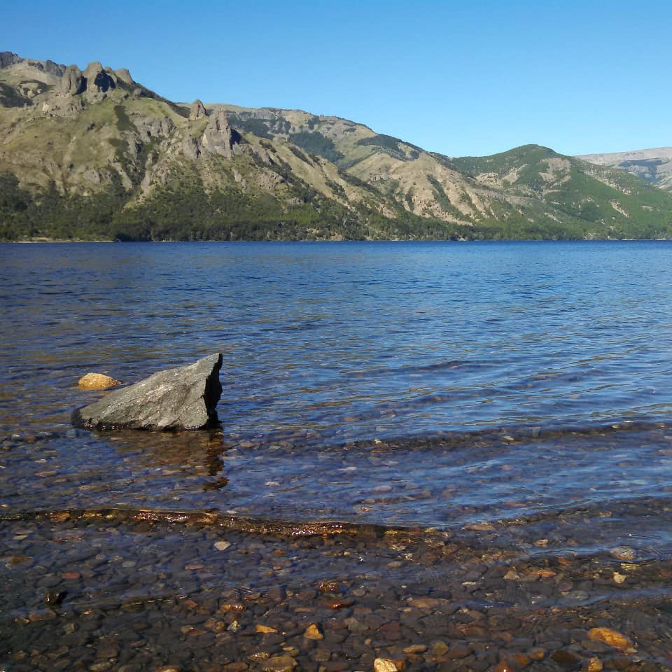 Vista desde la costa del lago, agua transparente y sierras de fondo.